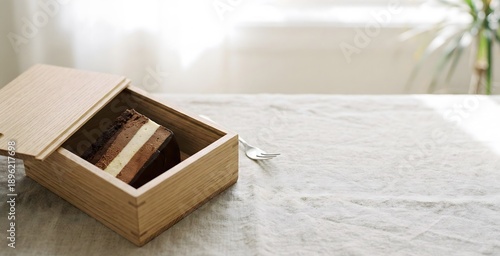 A delicate multi-layered chocolate and cream cake is presented in a natural wooden box on a textured table, ready to be enjoyed.