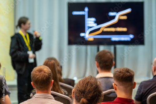Presenter leads conference session with audience watching large screen showing chart. Clear professional setting with focus on data visualization and learning