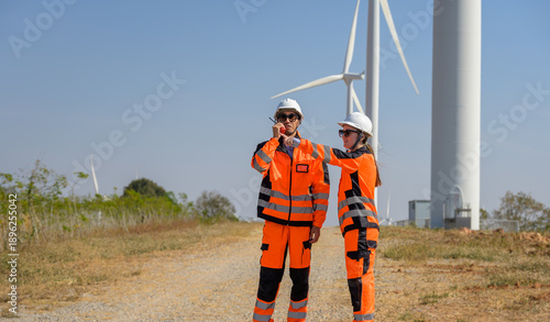 Two wind energy technicians wearing protective gear stand at wind farm and communicating with walkie talkie while inspecting turbines renewable power facility