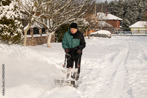 A senior man clears snow near his house. A person works with a snowplow. There is a lot of snow; winter, housing estate.