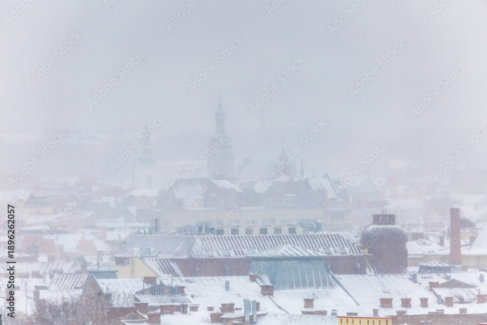 Fototapeta premium Distant view of Bernardine Monastery and St. Andrew's Church in Lviv during winter snowstorm. Scenic urban landscape of old town landmarks through thick fog and snowfall on cold January day.