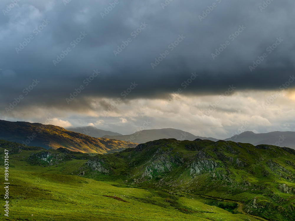 Fototapeta premium Rolling hills under a dramatic, cloudy sky. Green valleys and distant mountain peaks.