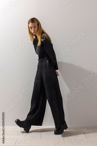 Schilderij op canvas Editorial fashion model walking forward in modern black jumpsuit, posing confidently in minimalist studio