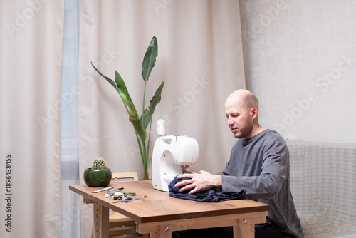 A bald man of 40 years old in a gray T-shirt sits at a table and sews on a sewing machine at home
