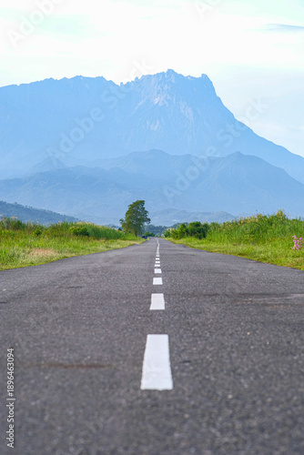 A low-angle shot of a straight paved road leading toward the majestic peaks of Mount Kinabalu under a clear sky in Sabah, Malaysia.