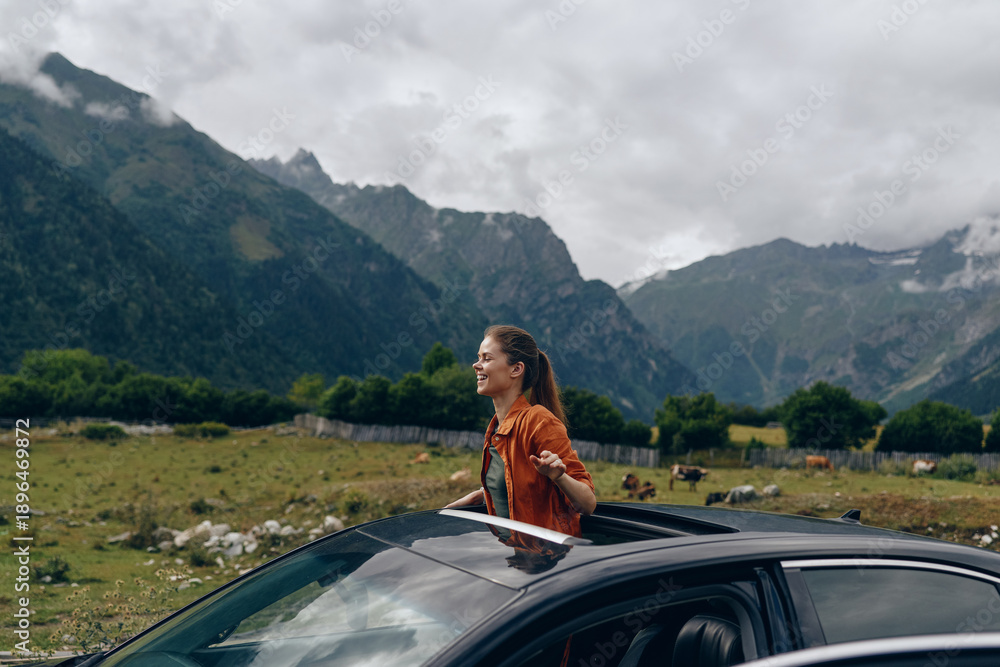 Obraz premium A woman stands beside a sleek car in a dramatic mountain landscape, enjoying the breeze and wide open scenery during a scenic road trip