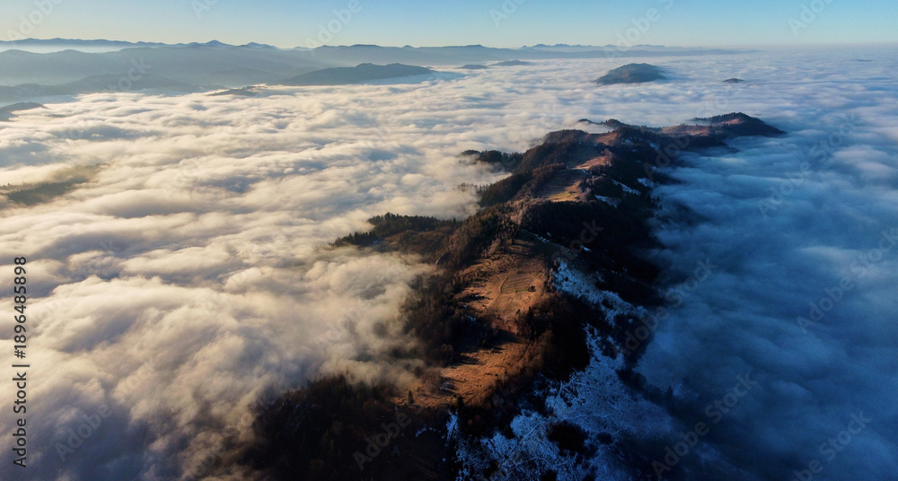 Fototapeta premium Breathtaking aerial view of mountain ridge piercing through sea of clouds. Sunlight illuminates patches of ridge. Distant mountain ranges create serene and expansive horizon.