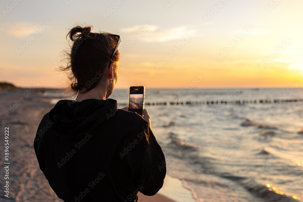 Fototapeta premium Woman photographing sunset seascape smartphone sandy beach enjoying calm summer evening travel lifestyle leisure moment technology use. Golden hour light sea waves horizon outdoor relaxation scene