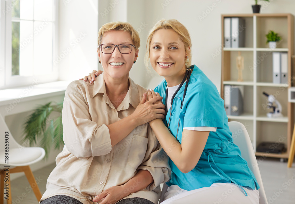 Obraz premium Smiling elderly woman sitting beside cheerful female caregiver in medical scrubs, holding hands with warmth in modern healthcare office. Concept of senior support, trust and professional care 