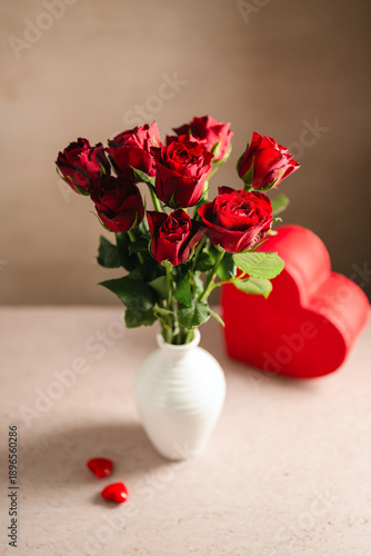 Valentine's Day festive background. Red roses flowers in a vase and heart gift box on neutral background. Festive greeting card, flat lay
