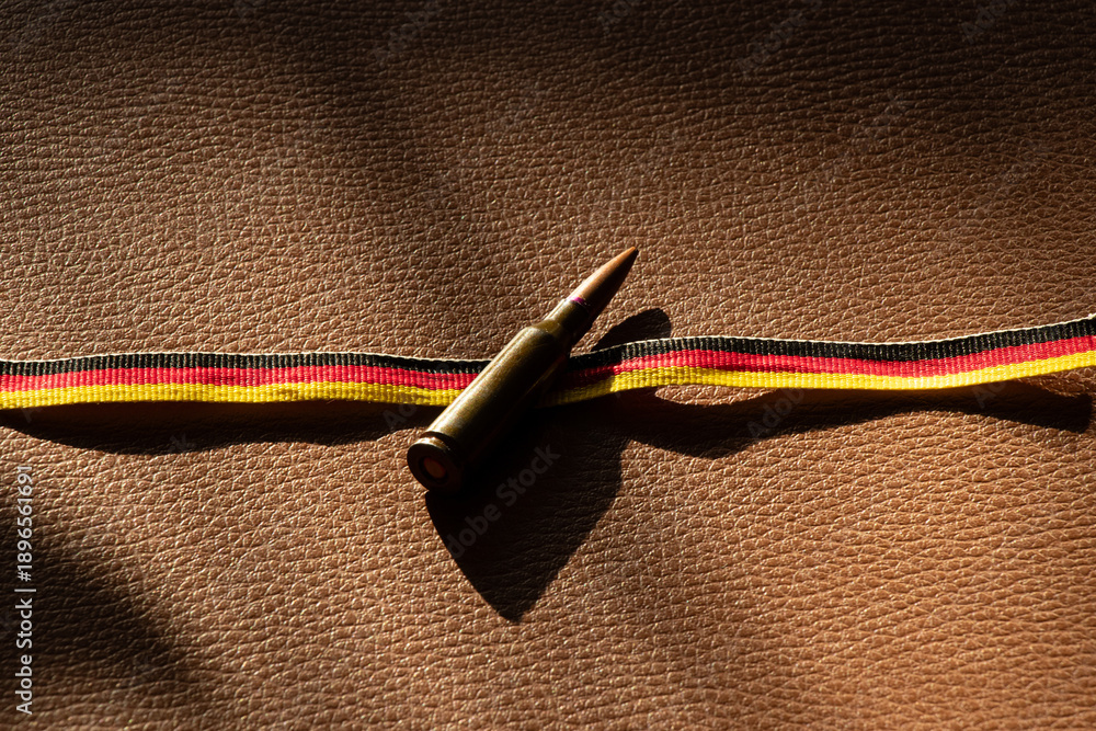 Fototapeta premium A bullet rests on a ribbon in the colors of the German flag on brown leather for themes of defense, army, weapons, security