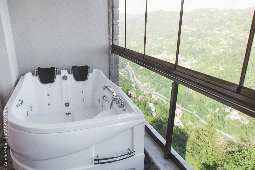 Fototapeta premium Modern white jacuzzi bathtub placed on a glass enclosed balcony with panoramic mountain valley view, symbolizing luxury wellness, spa relaxation, travel comfort and peaceful nature retreat lifestyle 