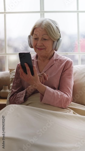 Glad senior woman in headphones listening to music in bed while using phone
