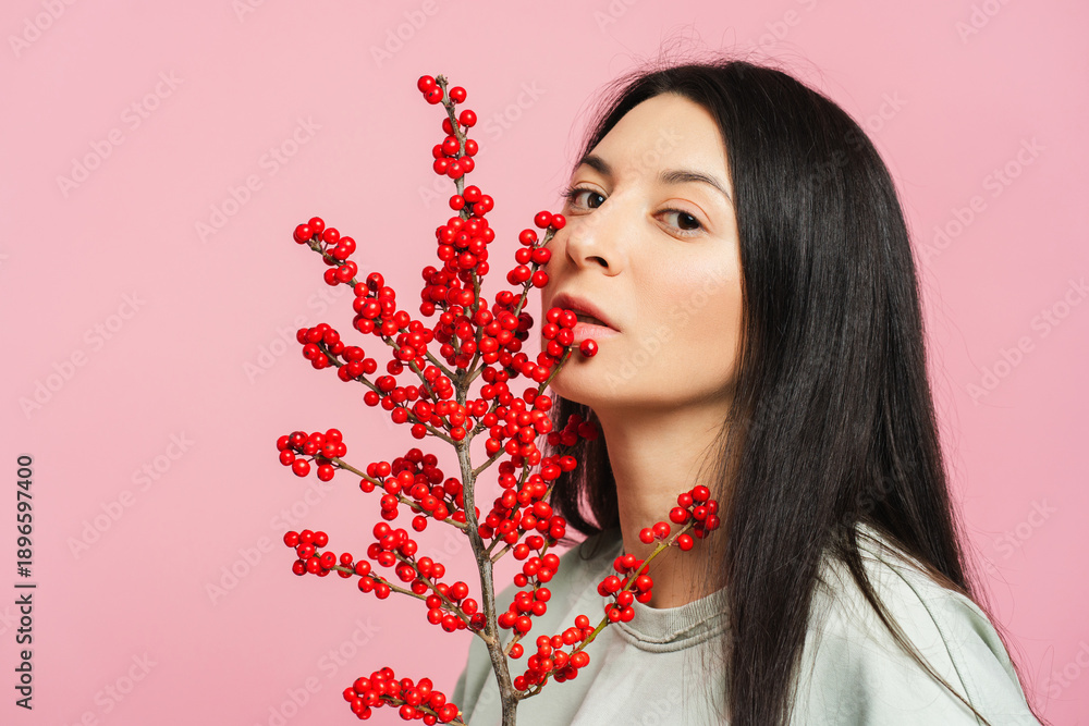 Fototapeta premium Serious pensive young woman holding twig with red berries on pink background. Birthday concept