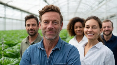 Group of five people standing in a greenhouse. the man in the center is a middle-aged man with short curly hair and a beard, wearing a blue denim shirt.