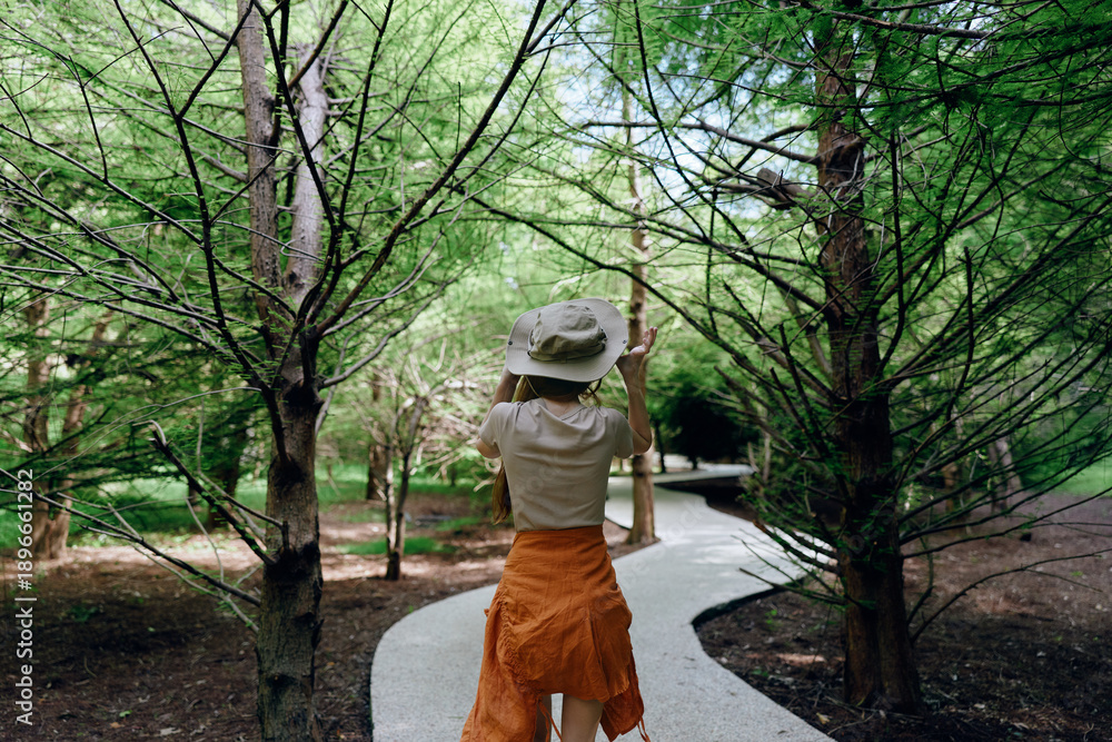 Obraz premium Woman walking on winding path through forest wearing hat and orange dress, nature trail with trees and dappled sunlight, casual summer stroll and peaceful outdoor exploration.