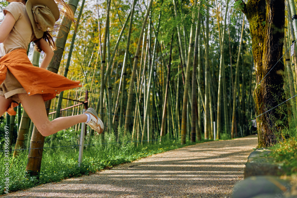 Obraz premium Girl jump on pathway in bamboo park with sun hat and orange skirt, summer motion captured midair, carefree child enjoying gravel trail under tall green bamboo trees and sunlight.