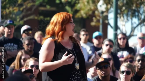 Redhead woman speaks with gestures to a crowd of people outdoors on a sunny day