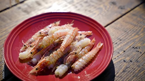 Raw shrimp and white pieces piled on a red plate on a rustic wooden surface, with a bottle drizzling liquid