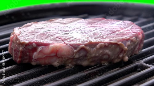 Raw, thick-cut steak grilling on a black, hot grill with a green background, steam & liquid pools on top