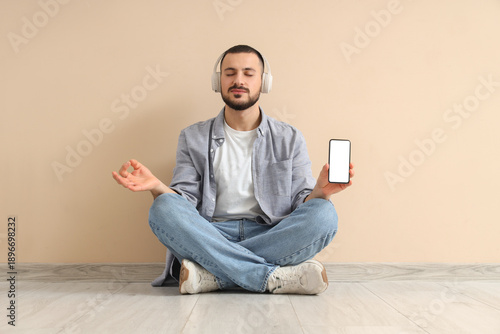 Handsome young man in headphones with mobile phone meditating near beige wall