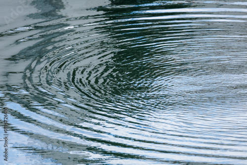 фотографія Close-up concentric ripples expanding in circular pattern across cool blue-green water surface partially reflecting daylight sky