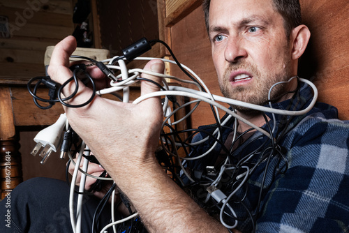 Cuadro en lienzo A stressed adult male struggles with a chaotic tangle of various electronic cables, USB cords, and chargers in a dark room
