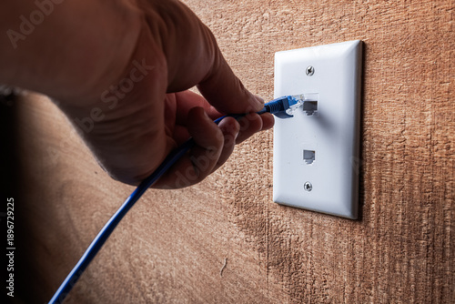 A close-up, high-detail shot of a hand plugging a blue RJ45 Ethernet cable into a white wall-mounted network port.