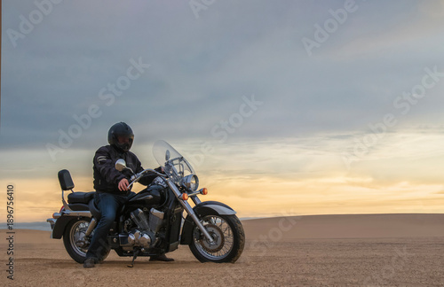 Exclusive Image: Off-road motorcyclist riding through the vast Namib Desert at golden hour, framed by sweeping sand dunes and a dramatic cloudy sky. A powerful scene of freedom, adventure travel