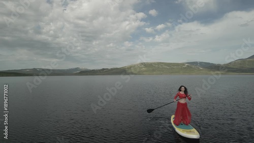 Raw drone footage flies directly toward a woman in a dress standing on a paddleboard on a mountain lake, then executes a smooth dolly-out reverse shot, backlighting her with sun rays.