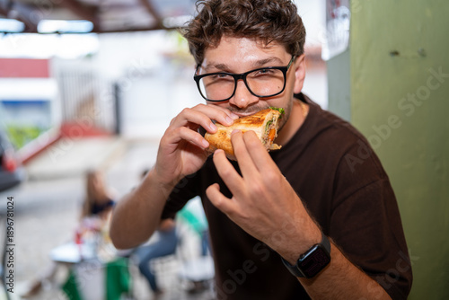 Young man eating sandwich at outdoor cafe, casual lifestyle food moment