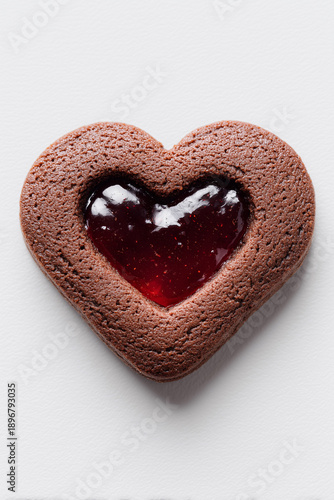 A delicious heart-shaped chocolate cookie with a glossy red jam center, photographed from a top-down perspective on a clean white textured background.