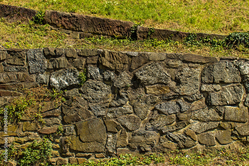 Fotografie Old stone retaining wall in a public park with green grass and trees on a sunny