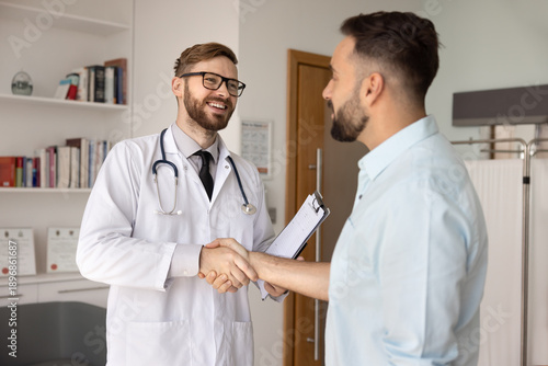 Successful doctor and patient interaction. Positive friendly young physician share firm handshake with male visitor in modern medical office start appointment with warm greeting show empathy respect