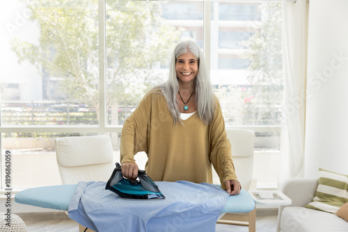 Fotografie Portrait of happy satisfied housekeeper or housewife posing in modern living living room stand at ironing board, smoothing-out shirt, smile, looking at camera