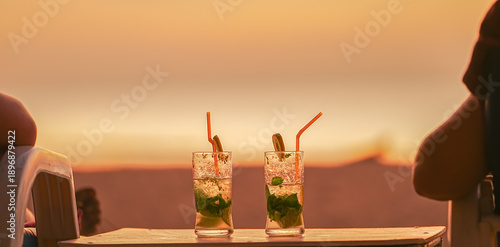 Two mojito cocktails in glass glasses with sunset background close-up. A couple in love are admiring the orange sunset on the beach. Travel, vacation and honeymoon resorts concept. High quality photo