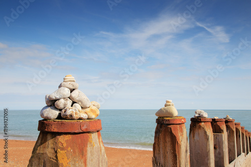Stacked Stone Cairns On Rusty Seawall Posts Along A Calm Beach With Blue Ocean Sky
