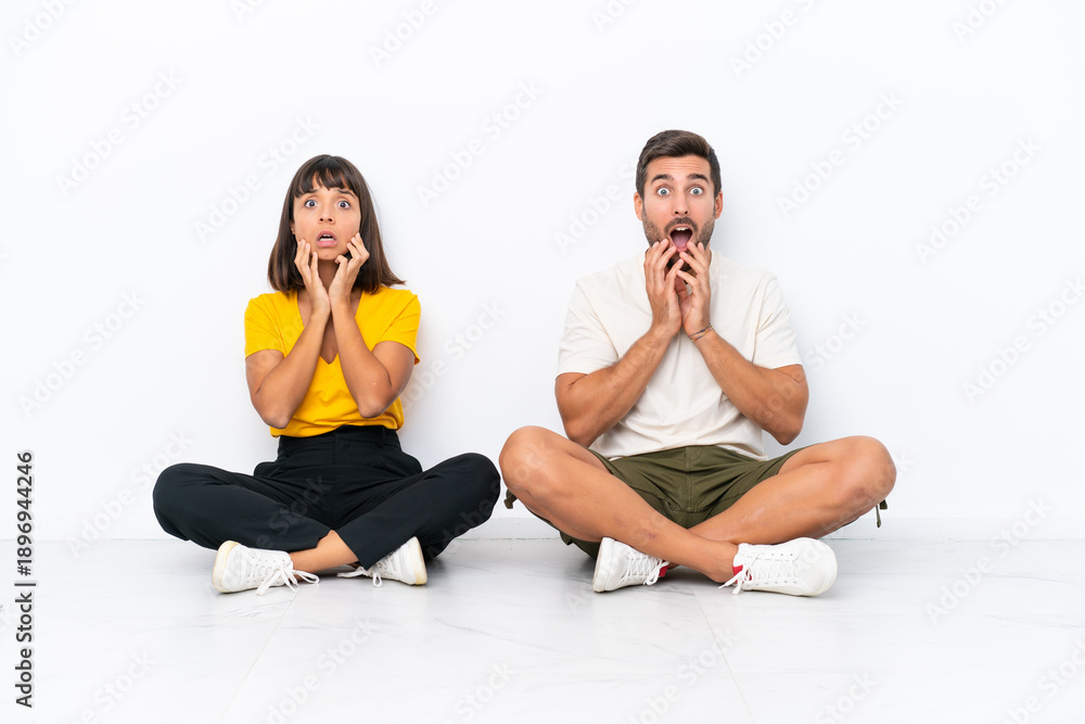 © luismolinero - Young couple sitting on the floor isolated on white background surprised and shocked while looking right