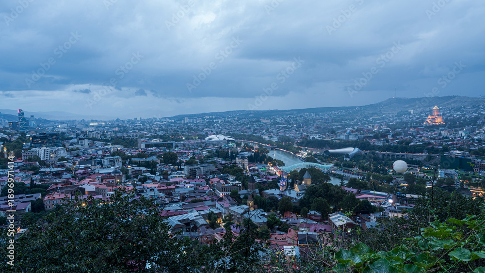 Obraz premium Evening cityscape of Tbilisi, Georgia, with cloudy sky and city lights reflecting on Kura River.