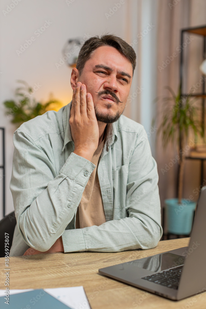 Naklejka premium Caucasian young man freelancer sitting at home office table feeling toothache touching mouth with hand, painful expression because of dental illness. gum disease, dental disease concepts. Vertical.
