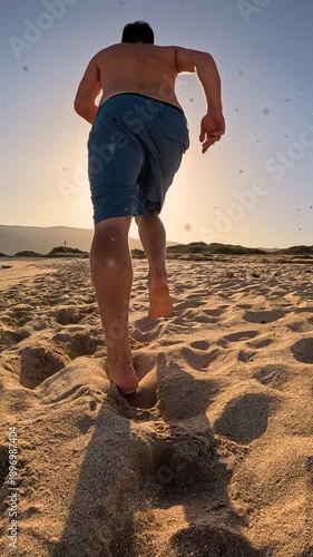 VERTICAL, LOW ANGLE VIEW, LENS FLARE: Fit young man in blue short during a morning run along sandy beach at Lanzarote. Disciplined sporty guy sticks to his healthy habits even on his summer vacation.