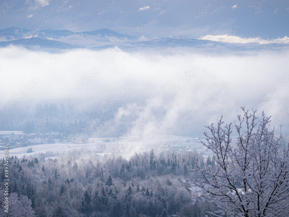 Fototapeta premium Remains of snowstorm clouds rolling over forested hilly countryside and villages