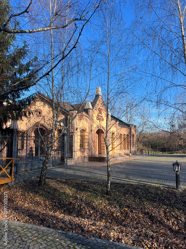 Historic brick building with arched windows and decorative details, set in a quiet park and framed by bare birch trees