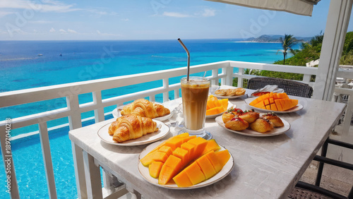 A tropical breakfast spread is placed on a balcony table. Fresh papaya and mango sit beside pastries. An iced latte is ready to drink. The ocean and resort pool are visible below in bright sunlight
