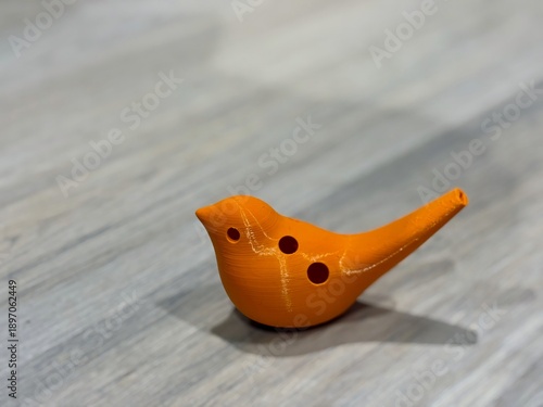 Orange 3D printed bird ocarina whistle on gray wooden floor, close-up handmade plastic musical instrument prototype with visible layer lines, DIY maker fabrication