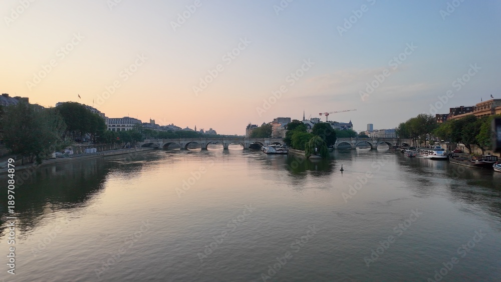 Fototapeta premium Seine river reflecting soft sunset colors with historic pont neuf arches and parisian buildings along tranquil urban banks at dusk, showing city life