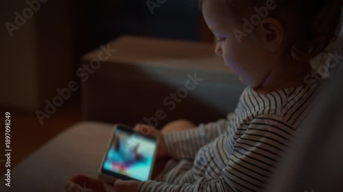 Young child snuggled up on the sofa is completely absorbed in vibrant cartoons on smartphone screen, raising concerns about gadget addiction and potential harm to their eyes and health in the evening