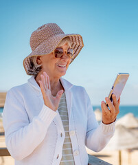 Video call concept. Smiling senior woman wearing hat and eyeglasses standing at the beach using mobile phone technology for online webcam connection - horizon over sea and blue sky