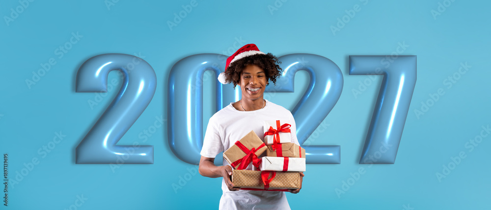 Fototapeta premium A person stands with a basket of various gifts, wearing a red festive hat. Behind them is a blue background with large numbers indicating the year ahead.