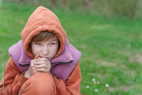 A boy in an orange hoodie sits on green grass on a cloudy spring day, dreaming about the future.